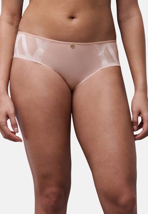 Woman wearing light pink hipster-style underwear with sheer geometric side panels and a small round gold charm at the waistband center.
