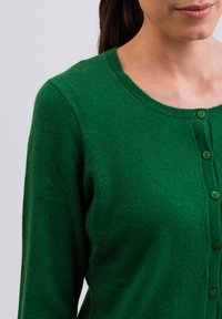 Grön cardigan med rund halsringning, gjord av mjukt tyg, med knappstängning framtill och ribbade muddar och fåll.