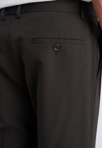 Primer plano de pantalones de vestir marrón oscuro con un bolsillo trasero abotonado y un bolsillo lateral en una persona que lleva una camisa blanca.