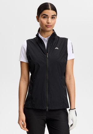 Kvinde iført en sort vest med lynlås over en hvid polo, sorte bukser og en hvid golfhandske på venstre hånd, stående mod en ensfarvet baggrund.