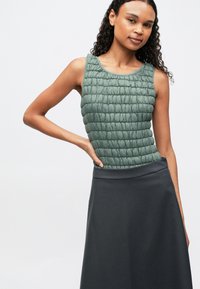 Moteris su garbanotais plaukais vilki be rankovių žalią pūkinį topą ir tamsią A formos sijoną, pozavo su viena ranka ant klubo prieš vientisą foną.