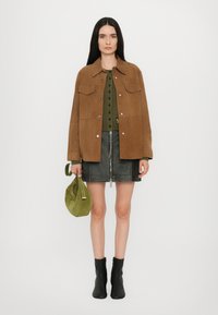 Giacca in suede marrone sopra una blusa verde con bottoni, abbinata a una gonna mini in pelle nera e stivaletti neri. Tieni una borsa verde.
