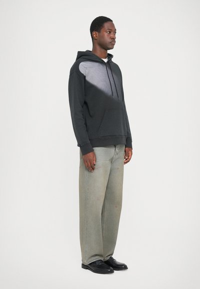 Črna kapuca z sivo gradientno barvo spredaj, kenguru žep, v kombinaciji s svetlo sivo širokimi hlačami in črnimi čevlji.