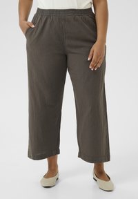 Personne portant un pantalon court ample marron foncé avec taille élastique et poches, associé à des chaussures plates beiges sur un fond blanc.