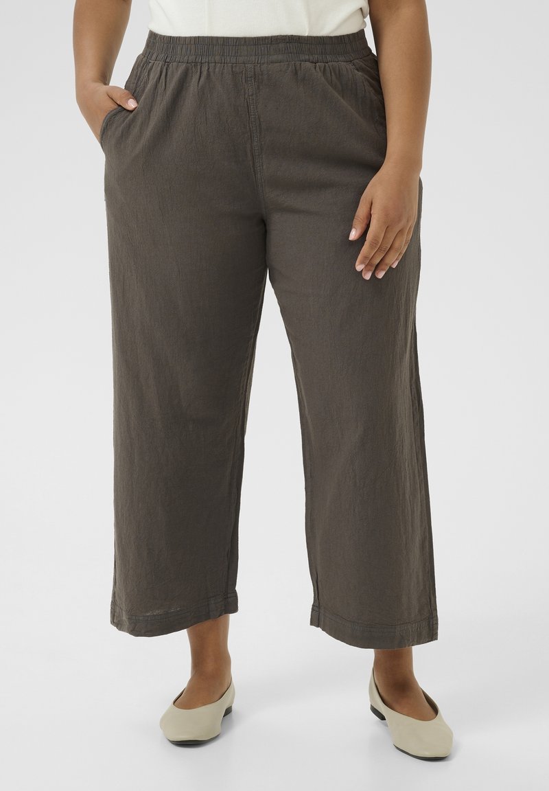 Personne portant un pantalon court ample marron foncé avec taille élastique et poches, associé à des chaussures plates beiges sur un fond blanc.