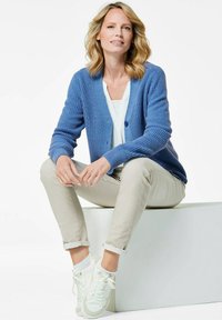 Gilet tricoté bleu clair avec texture côtelée, deux boutons, associé à un haut blanc et un chino beige retroussé ; des baskets blanches complètent le look.