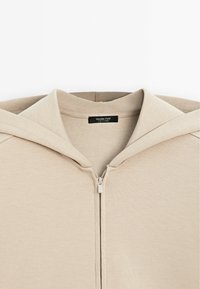 Haut à zip beige avec une grande capuche, texture de tissu douce et une étiquette à l'intérieur indiquant "Massimo Dutti". Design simple et minimal.