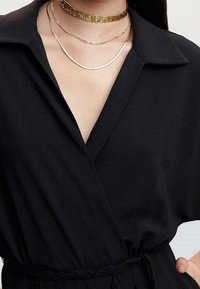 Žena v černých zavinovacích šatech s límečkem, s pleteným opaskem a třemi zlatými náhrdelníky, včetně chokeru s texturovaným designem.