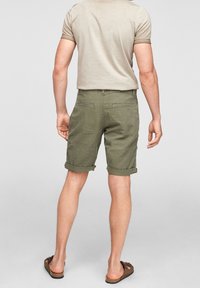 Shorts vert olive à coupe décontractée, ourlets roulés et deux poches arrière. Portés avec une chemise à manches courtes beige clair et des sandales.