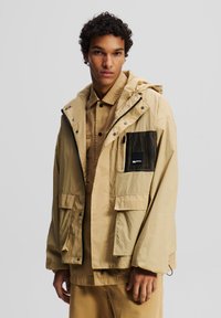 Beige Kapuzenjacke mit einem Frontreißverschluss, mehreren Taschen, einer Netz-Tasche auf der Brust und einem lässigen Schnitt, kombiniert mit einem passenden Shirt.