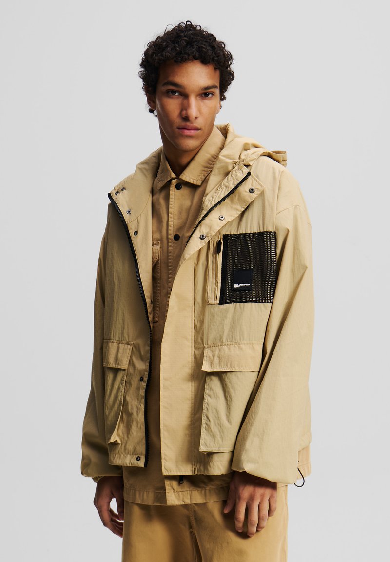 Beige Kapuzenjacke mit einem Frontreißverschluss, mehreren Taschen, einer Netz-Tasche auf der Brust und einem lässigen Schnitt, kombiniert mit einem passenden Shirt.