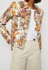 Giacca di jeans con motivo floreale nei colori vivaci di giallo, arancione e rosa, abbinata a una maglietta bianca e pantaloni beige chiaro, con chiusura a bottoni.