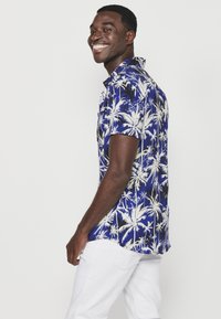 Camicia a maniche corte con bottoni, sfondo blu e stampa di palme bianche. Presenta una vestibilità rilassata e colletto.