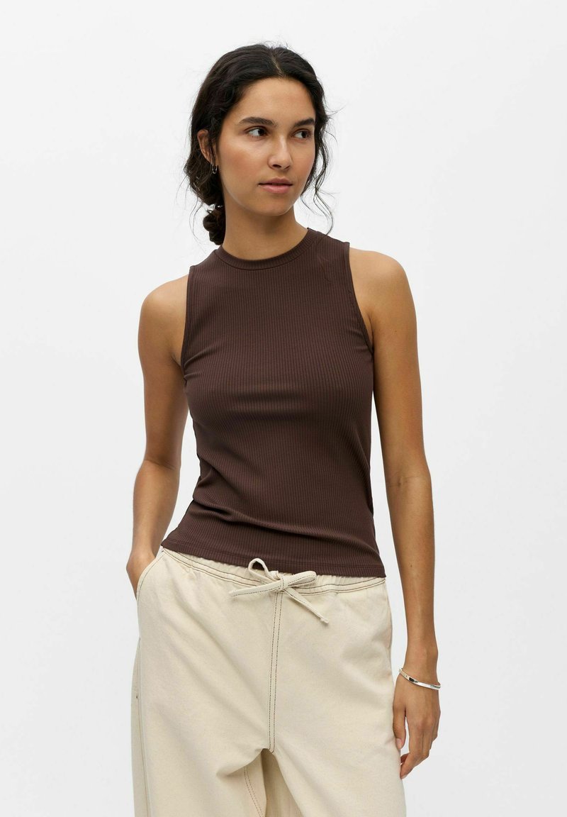 Vrouw met donker haar in een lage knot, draagt een mouwloos bruin geribd topje en lichtbeige broek met aantrekkoord tegen een effen achtergrond.