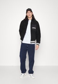 Sort fleecejakke med lynlås og lomme, prydet med hvid 'Champion'-logo; sat sammen med en lys skjorte, løse jeans og hvide sneakers.