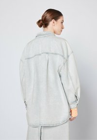 Giacca di jeans oversize azzurro chiaro con orlo arrotondato, texture sbiadita e cuciture visibili. Presenta un yoke posteriore e polsini delle maniche con zip.