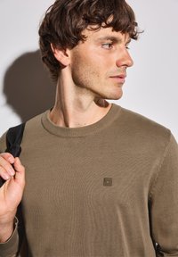 Olivgrön stickad tröja med rund halsringning, texturerad tyg och en liten logotyp på bröstet; modell som håller i ett svart band.