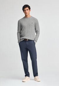 Grauer Cable-Knit-Pullover mit rundem Ausschnitt, kombiniert mit dunkelblauen Chinos und beigen Sneakers. Auffälliges Markenlogo auf dem Pullover.