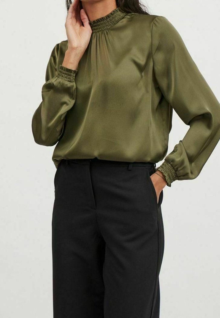 Femme portant un chemisier en satin vert olive à manches longues avec col et poignets froncés, associé à un pantalon noir taille haute.