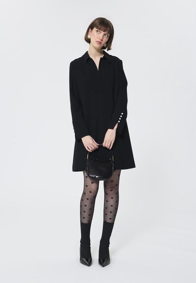 Schwarzes Langarmkleid mit Kragen, dekorativen Knöpfen an den Ärmeln, kombiniert mit schwarzen Strumpfhosen mit Herzmotiv und einer kleinen, glänzenden Handtasche.