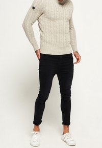 Beżowy sweter w splot warkoczowy o prążkowanej fakturze, zestawiony z dopasowanymi czarnymi jeansami i białymi tenisówkami. Charakterystyczny czarny akcent na rękawie swetra.