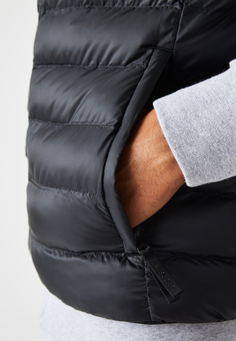 Le gilet rembourré noir présente des sections matelassées, une poche latérale zippée et une texture lisse et élégante, associé à une manche grise en dessous.