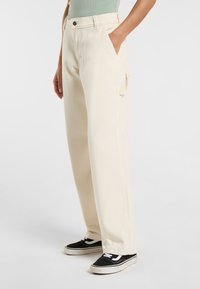 Pantaloni a gamba larga color crema realizzati in tessuto morbido, con tasche laterali e chiusura con bottone, abbinati a sneakers nere.