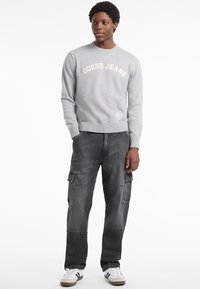 Sudadera gris de cuello redondo con el logo "GUESS JEANS", combinada con pantalones cargo grises que tienen bolsillos laterales y zapatillas blancas. Textura suave.