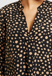 Blusa nera con scollatura a V profonda, caratterizzata da grandi pois beige e un materiale morbido e leggero. Il tessuto ha una leggera lucentezza e cade delicatamente.