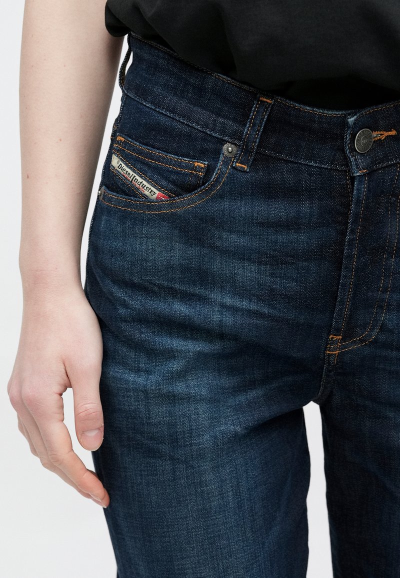 Vaqueros de mezclilla oscura con costuras naranjas, diseño de talle alto y una etiqueta de marca visible en el bolsillo delantero. Textura suave y ajuste entallado.