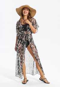 Kimono noir et blanc transparent à motifs, avec taille à nouer, manches longues et fentes latérales, porté par-dessus un maillot de bain noir et des tongs.