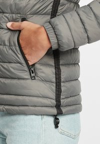 Giacca a vento grigia con zip laterale, polsini a costine e orlo regolabile tramite cordino, dal design elegante e leggero con superficie texture.