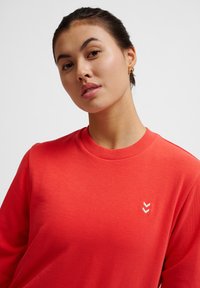 Czerwony sweter z okrągłym dekoltem, z małym białym logo w kształcie chevronu na lewej piersi. Gładki materiał, luźny krój i ribbed cuffs.