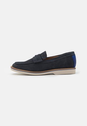 Mocassin en suede bleu marine avec un bout arrondi, une sangle décorative cousue sur le devant, et des accents bleus contrastants au niveau du talon. Semelle en caoutchouc.