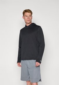 Svart hettegenser med lange ermer og teksturert overflate, sammen med lysegrå shorts. Har en liten hvit logo på venstre erme.