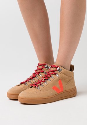 Tan-farbene Wildleder-Hochtop-Sneaker mit roten und schwarzen Schnürsenkeln sowie einem orangefarbenen "V"-Logo, getragen an nackten Beinen vor einem weißen Hintergrund.