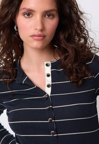 Haut à manches longues côtelé bleu marine avec des rayures horizontales blanches, doté d'une patte de boutonnage partielle avec des boutons noirs et d'un col blanc contrastant.