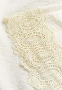 Tissu couleur crème avec un bord en dentelle ovale complexe comprenant un fin filet et des motifs de points décoratifs le long du bord.