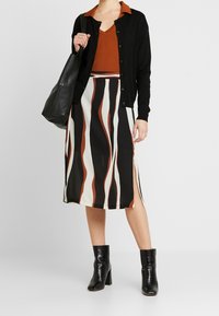 Svart cardigan över en orange ribbad topp, kombinerad med en knälång kjol med vågiga svarta, vita och rostiga mönster. Svarta ankelstövlar.