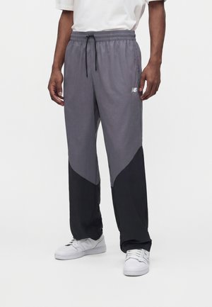 Uomo che indossa pantaloni della tuta New Balance grigi e neri a blocchi di colore con scarpe da ginnastica bianche e t-shirt bianca, in piedi su sfondo bianco.