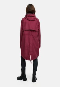 Bordowy płaszcz przeciwdeszczowy z kapturem, o asymetrycznym dolnym wykończeniu, z bocznymi kieszeniami i ściągaczem w talii, zestawiony z czarnymi skinny jeansami i botkami.