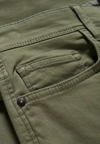 Olivgröna bomullsbukser med en framficka, detaljerad sömnad och en metalldetalj knapp. Tyget har en slät, lätt strukturerad yta.