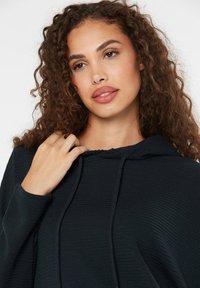 Schwarzer, gerippter Kapuzenpullover mit Kordelzug, der eine entspannte Passform und ein strukturiertes Material aufweist. Die Hand des Models ist in der Nähe des Halsausschnitts positioniert.