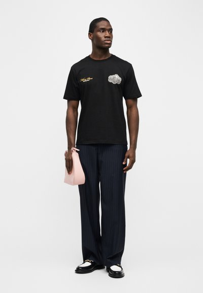 Άνδρας με μαύρο γραφικό μπλουζάκι, μπλε παντελόνι με ρίγες, μαύρα και άσπρα loafers, κρατώντας μια μικρή ροζ τσάντα χειρός σε λευκό φόντο.