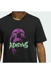 Schwarzes T-Shirt mit kurzen Ärmeln, das ein auffälliges lila Schädel-Graphic mit grütem Text zeigt, aus weichem Stoff und mit normaler Passform.
