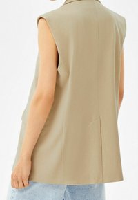 Beżowa bezrękawnik, dopasowany krój, gładka tekstura, wyrazisty kołnierz i delikatne wycięcie z tyłu. Połączona z jasnoniebieskimi dżinsami.