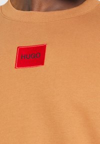 Khakifarbendes, geripptes T-Shirt mit einem rechteckigen roten Stoffpatch, auf dem "HUGO" in Schwarz gestickt ist, in der Nähe des Ausschnitts platziert.