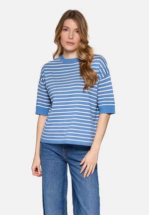 Joven con cabello largo y ondulado, vestida con un suéter de manga corta a rayas azul y blanco y jeans azules, de pie frente a un fondo liso.