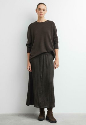 Frau trägt einen dunkelbraunen übergroßen Pullover, einen plissierten langen Rock und braune Knöchelstiefel und steht vor einer weißen Wand.