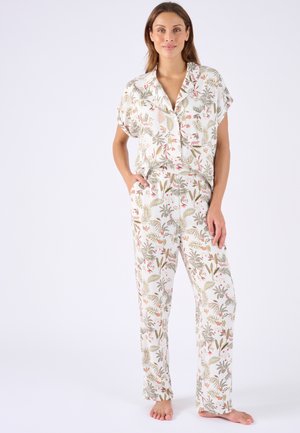 Frau steht barfuß da, trägt eine weiße, kurzärmelige Bluse mit Blumenmuster und passende Hose, Hände in den Taschen, vor einem schlichten Hintergrund.